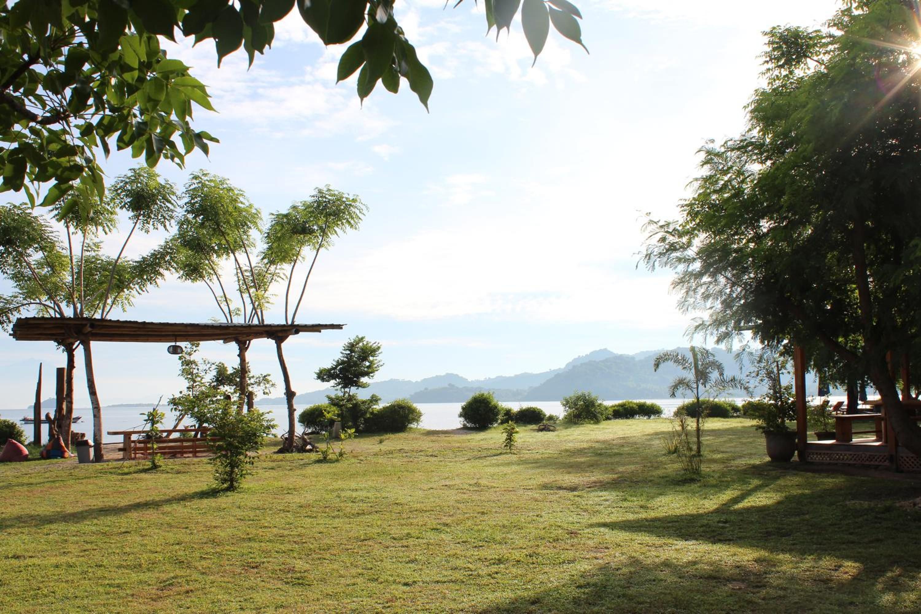 A grassy outdoor area with a wooden pergola, trees, and a view of a body of water with mountains in the distance.