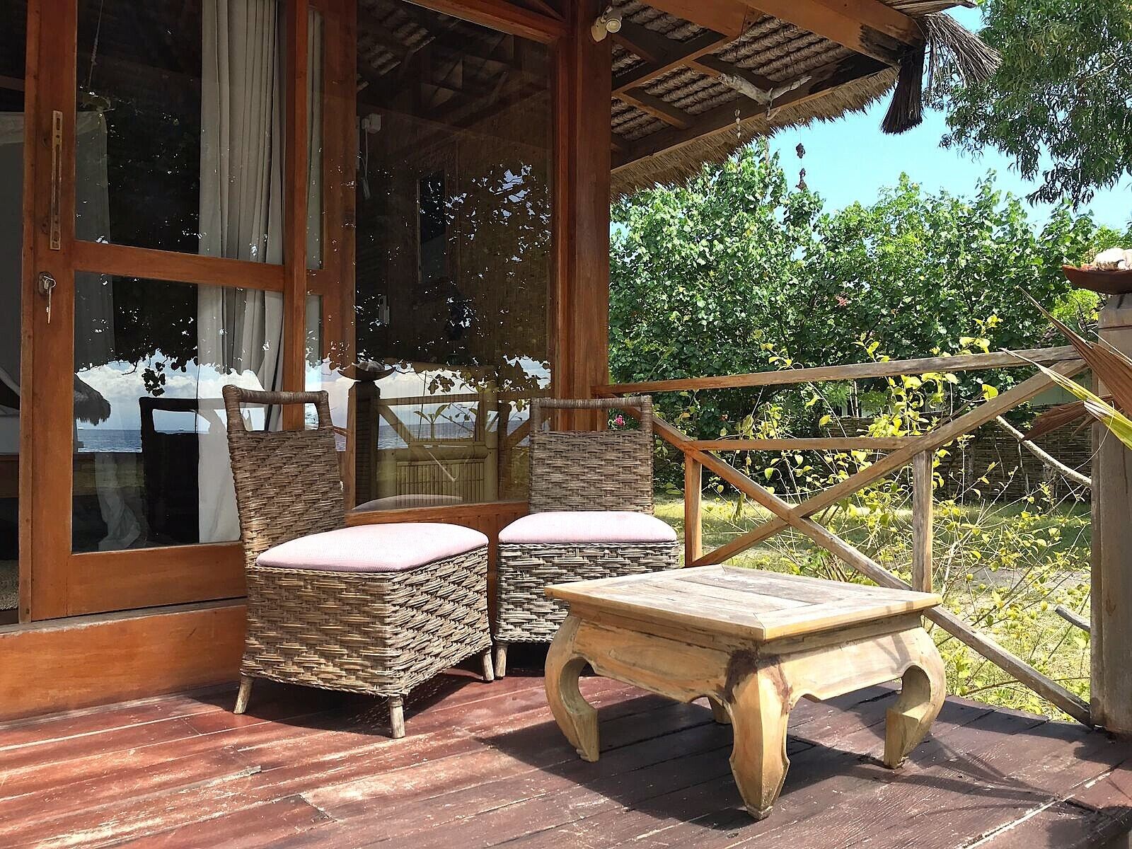A wooden deck with wicker chairs and a wooden table, overlooking a garden and a view of the sea.