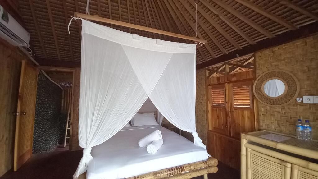 A room with a bed under a mosquito net, wooden walls, a round mirror, and a wooden cabinet with bottles.