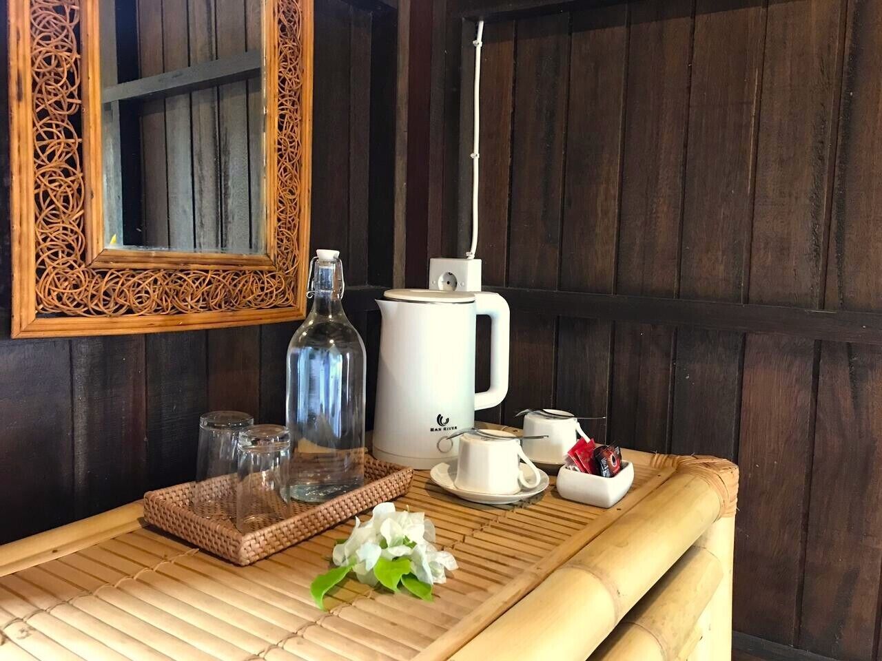 Una mesa de madera con una bandeja tejida, una botella de vidrio, una tetera eléctrica blanca, una taza y una pequeña flor blanca.