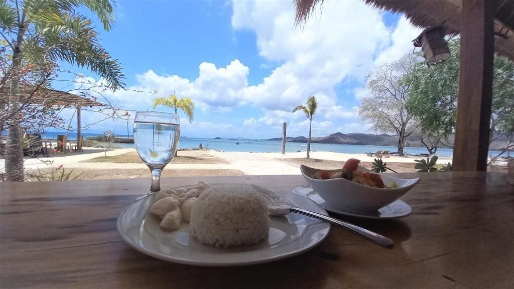 Uma mesa de jantar com um prato de arroz e uma tigela de comida, um copo de água, e vista para a praia e o oceano.
