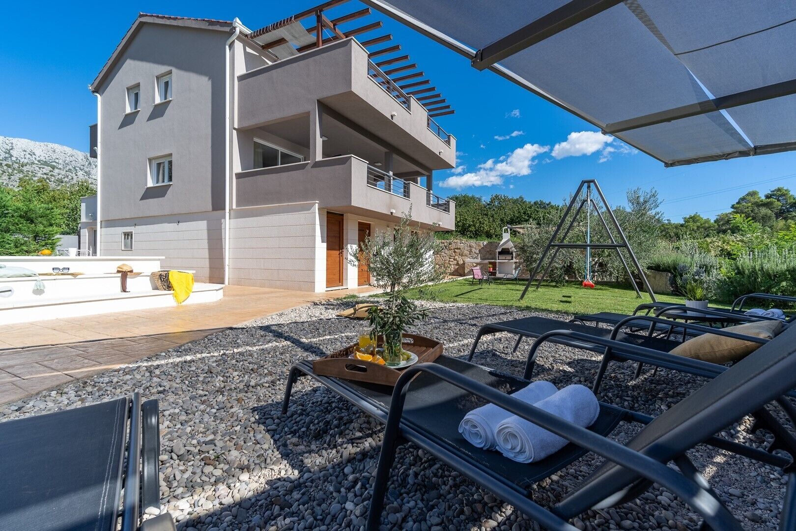 A modern two-story building with a balcony, outdoor seating area with lounge chairs, and a small garden.