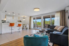 A modern living room with a sofa, ottoman, and two bar stools. There is a dining area with a table and chairs. The room has large windows and a balcony with a view of greenery. - Villa Jelena private pool no-Chlorine system,Sauna (Naklice)