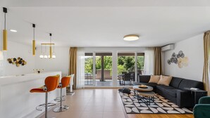 Ein modernes Wohnzimmer mit einer Couch, einem Couchtisch und orangenen Sesseln. Ein Balkon bietet Blick auf Grünflächen.