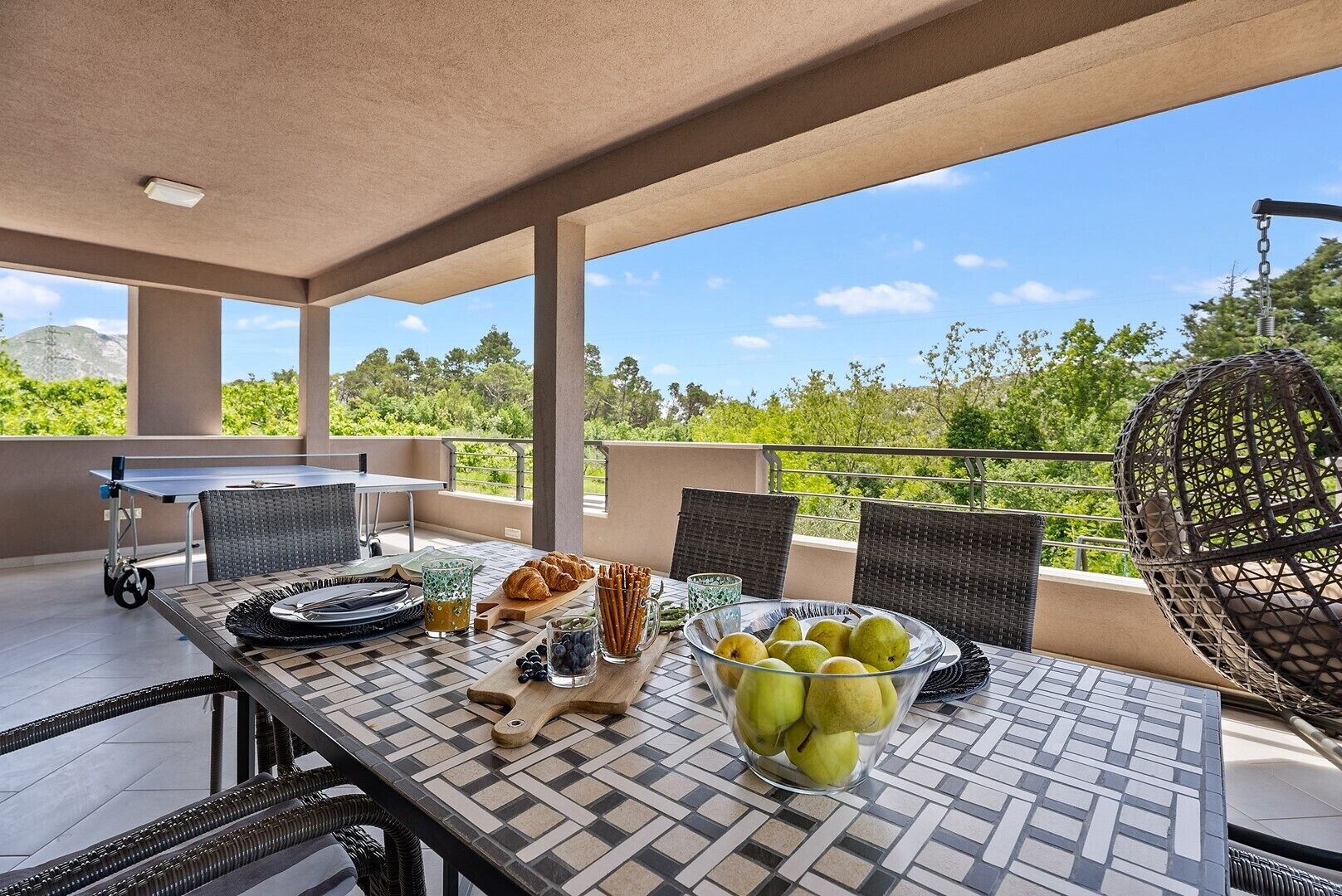 Um terraço com uma mesa posta para o pequeno-almoço, incluindo croissants, uma taça de fruta e uma bebida.