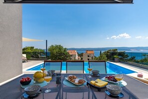 A dining table with breakfast items and drinks, overlooking a pool and a scenic view of the sea and buildings. - Villa Soriano with heated pool & sea view, 130m from sea, 8 persons max (Mimice)