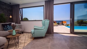 Ein modernes Wohnzimmer mit einer Couch, einem Türkis-farbenen Sessel und einem kleinen Tisch mit einer Vase. Große Fenster bieten Blick auf einen Pool und ein Gebäude gegenüber dem Wasser.