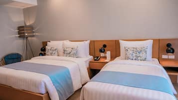Une chambre d’hôtel avec deux lits, chacun doté d’une tête de lit en bois, de draps blancs et d’un élément décoratif bleu. On y trouve une table de chevet avec une lampe, un téléphone et une petite lampe de table. Une lampe sur pied est visible dans un coin.
