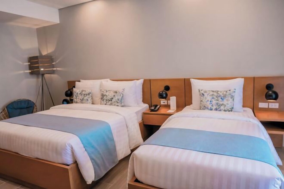 A hotel room with two beds, each with a wooden headboard, white bedding, and a blue accent. There is a bedside table with a lamp and a phone on each side.