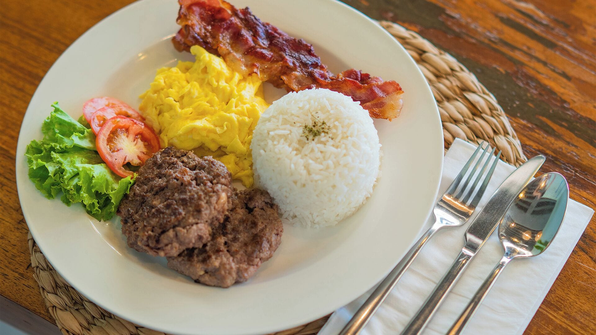 Un plato con huevos revueltos, tocino, arroz, una hamburguesa y una ensalada.