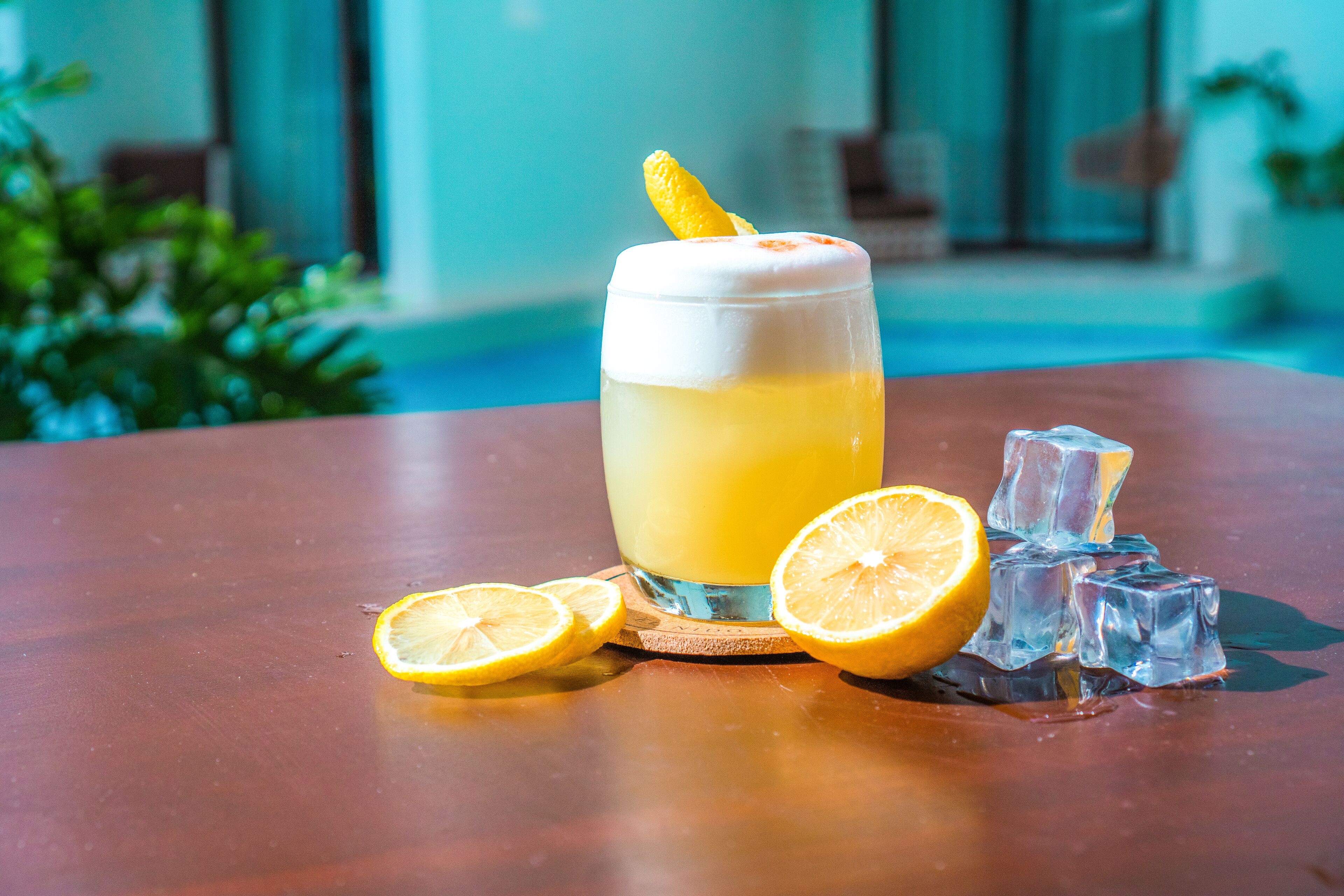 A glass of a yellow beverage with a lemon garnish, ice cubes, and lemon slices on a wooden table.
