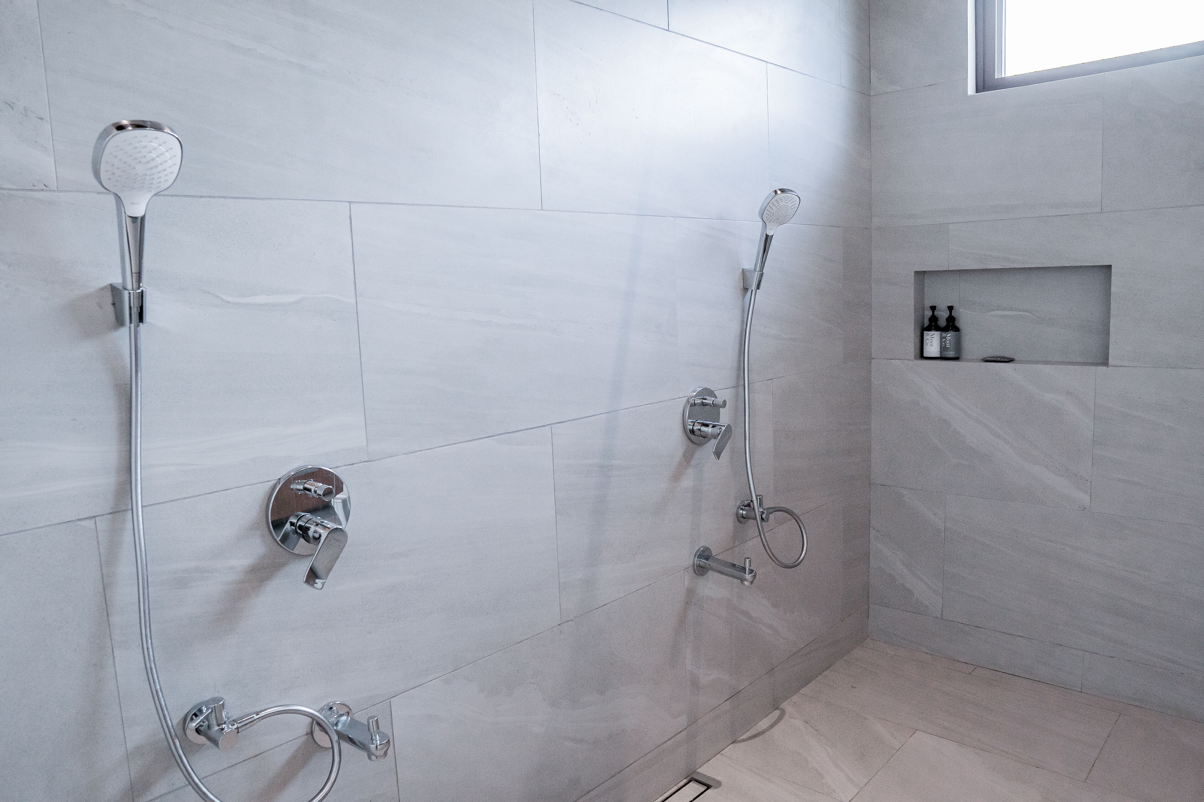 Un baño moderno con dos duchas, un estante empotrado con artículos de aseo y un ventanal para luz natural.