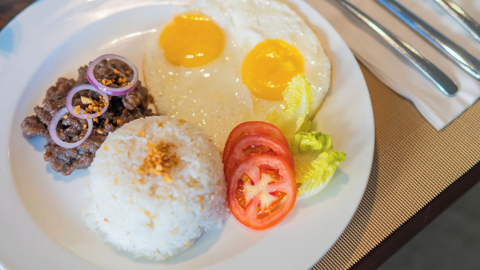 A plate with fried eggs, rice, a slice of tomato, lettuce, and a side of meat with onions and peanuts.