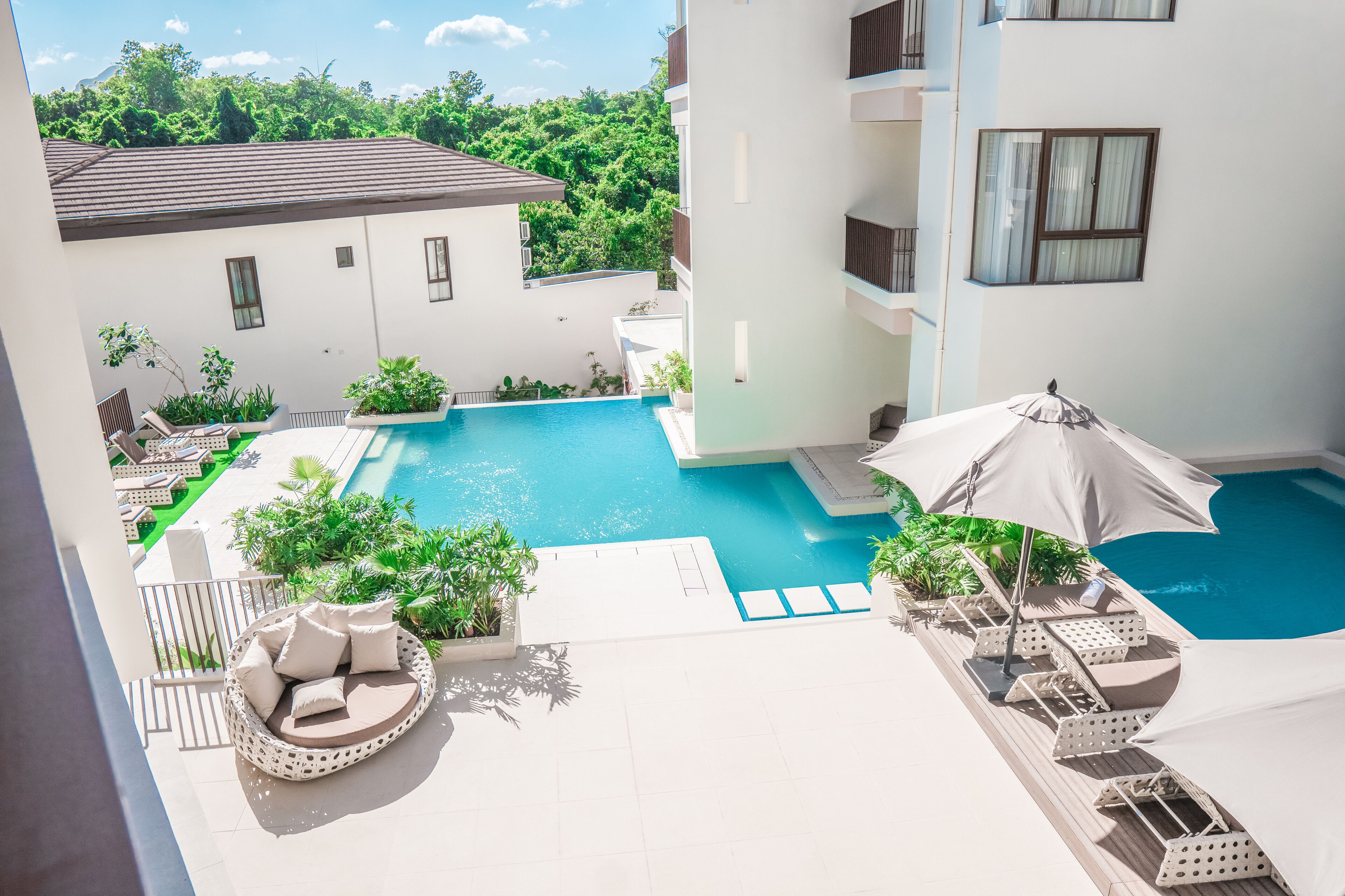A rooftop pool area with lounge chairs, umbrellas, and a view of surrounding greenery.