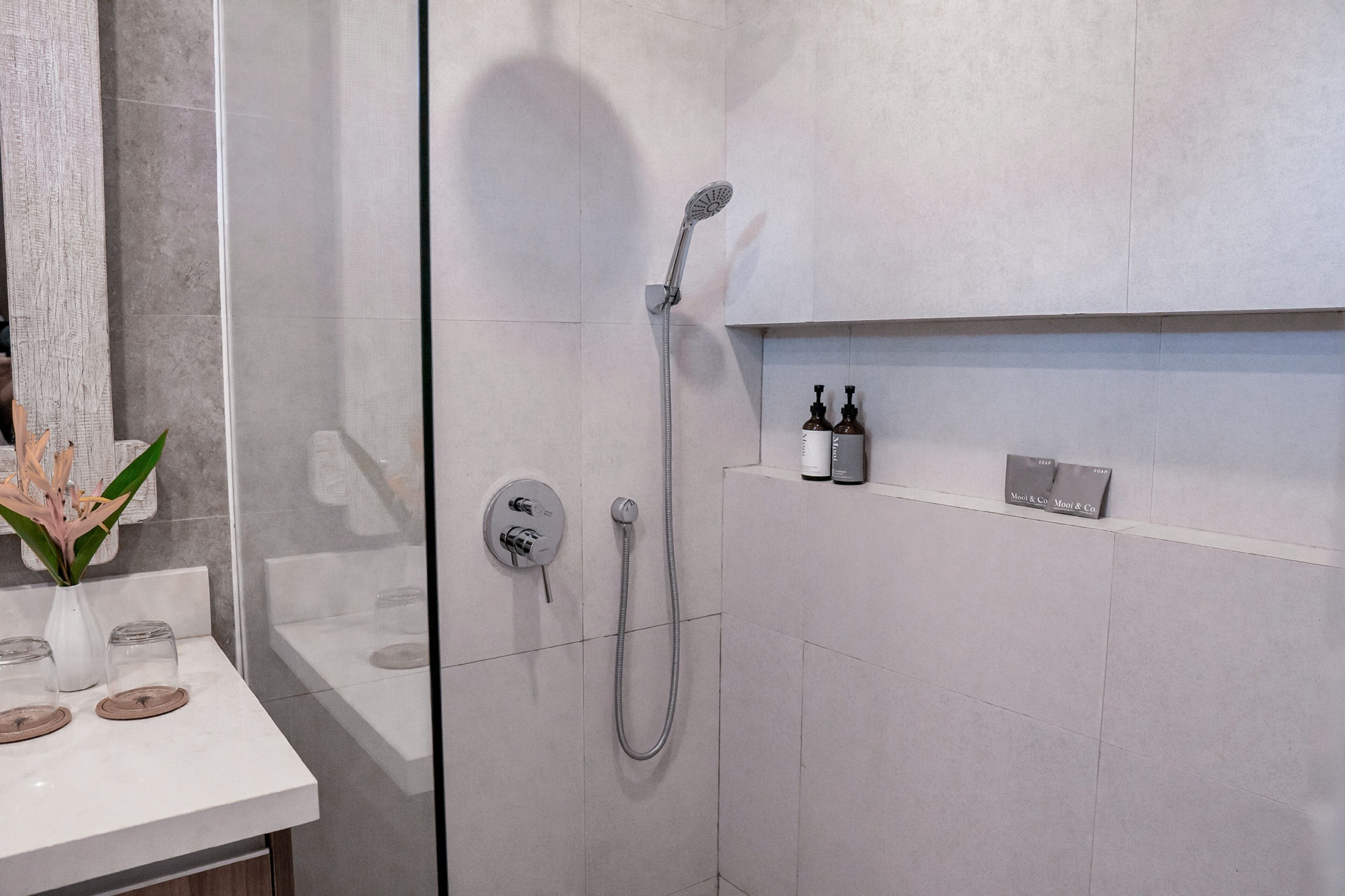 A modern bathroom with a glass shower enclosure, a white sink with a vase of flowers, and a shelf with toiletries.