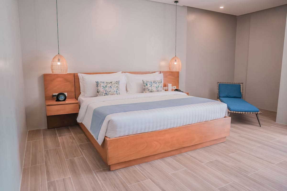 Un dormitorio moderno con una cama de madera, dos mesitas de noche, un buró, un sillón azul y dos lámparas colgantes.