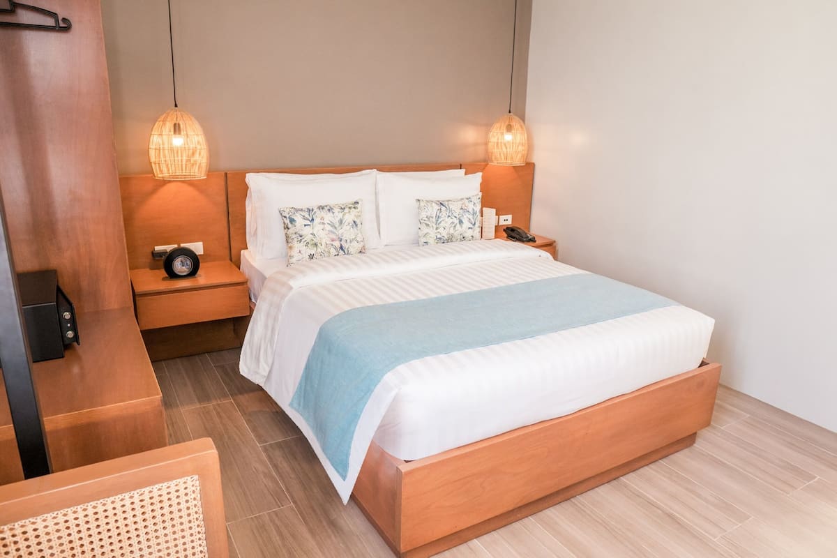 Habitación de hotel ordenada con una cama de madera, dos lámparas de noche, una mesita de noche y una pequeña mesa con un teléfono.