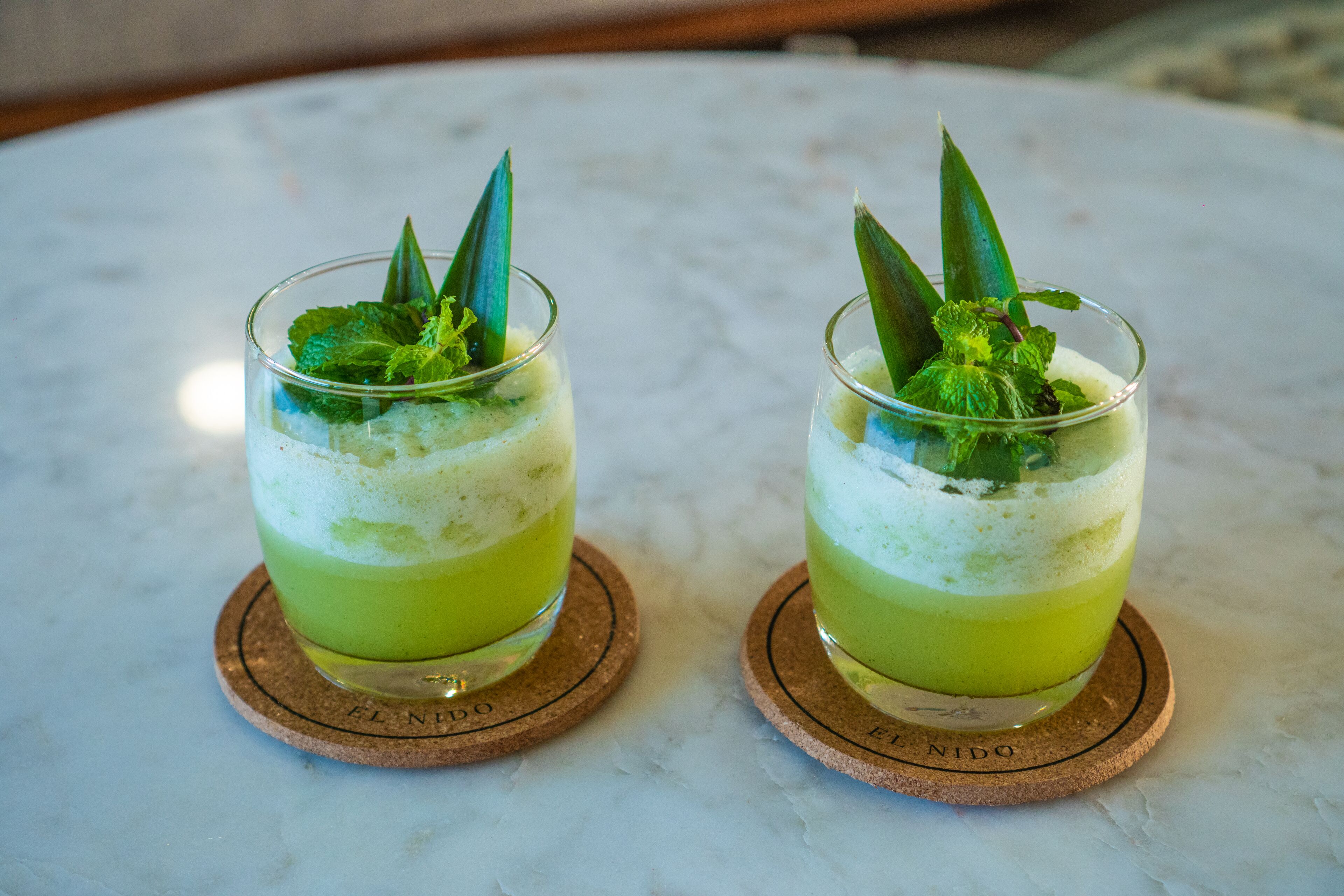 Two glasses of green beverage on a marble table with cork coasters.