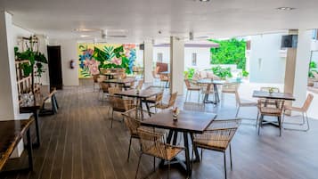 Uma ampla área de refeições interna com várias mesas e cadeiras de vime, apresentando um grande mural de flores tropicais na parede do fundo e vista para o verde lá fora.