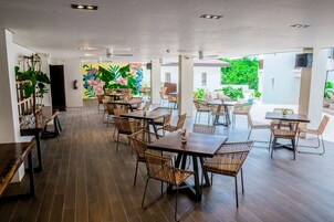 Uma ampla área de refeições interna com várias mesas e cadeiras de vime, apresentando um grande mural de flores tropicais na parede do fundo e vista para o verde lá fora.