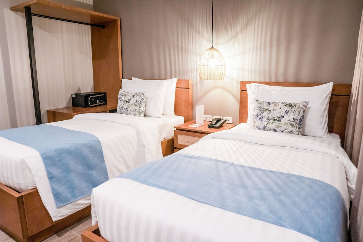 Una habitación de hotel con dos camas, cada una con cubrecama azul y blanco, una mesita de noche con lámpara y una radio pequeña.