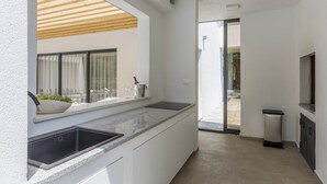 Una cocina moderna con un lavamanos integrado, una ventana con cortinas, y un pequeño espacio exterior con un contenedor de basura y un televisor.