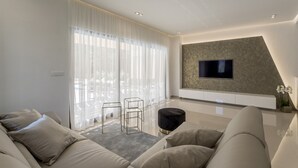 Una sala de estar moderna con un televisor de pantalla plana montado en una pared texturizada, un sofá, una mesa de centro y un ventanal con cortinas translúcidas.