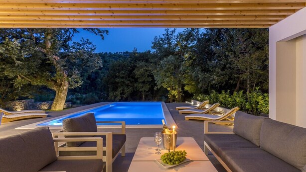 Un área de piscina moderna al aire libre con sillas reclinables y una mesa, rodeada de árboles.