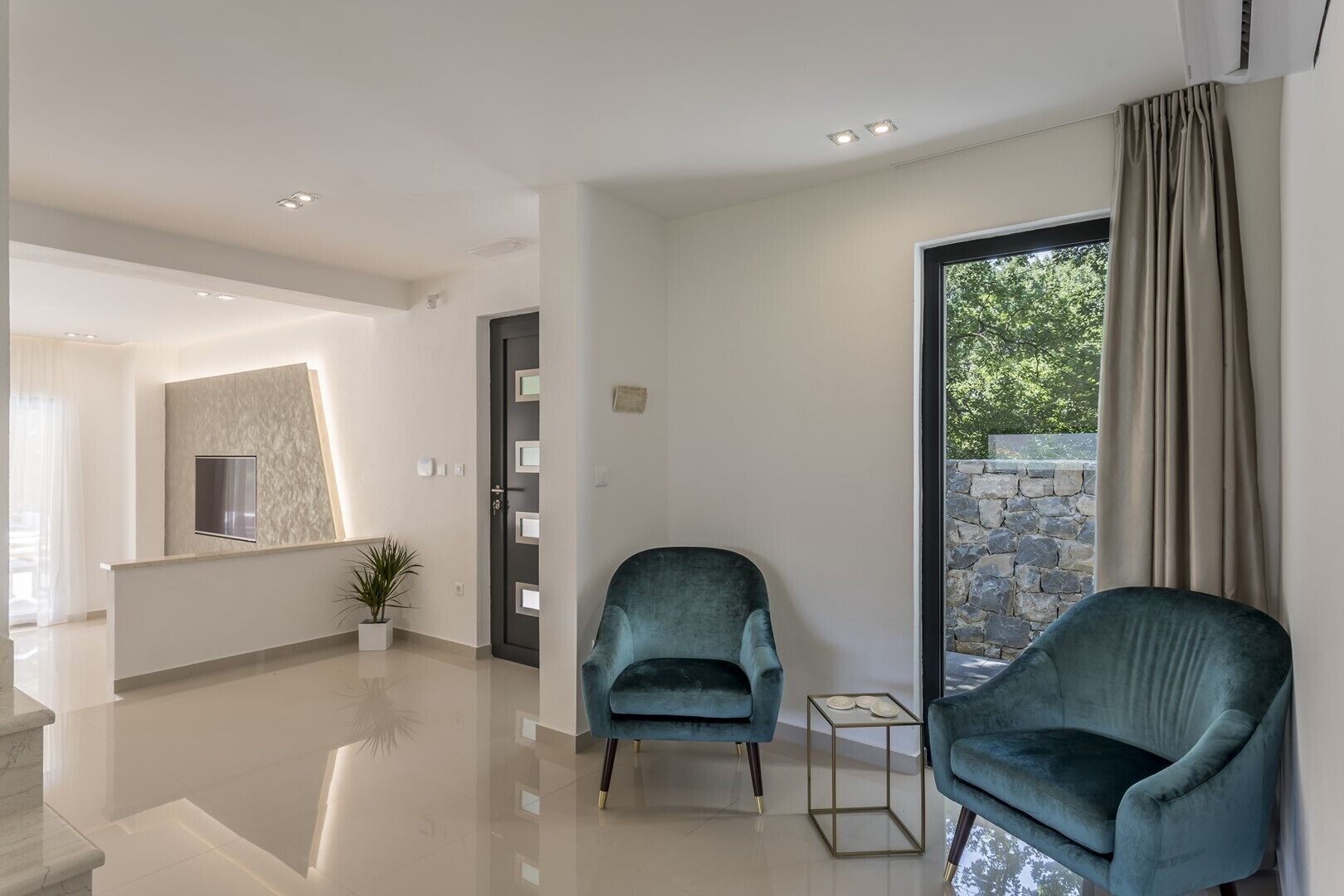 A modern living room with a large window, a stone wall, and two teal armchairs.
