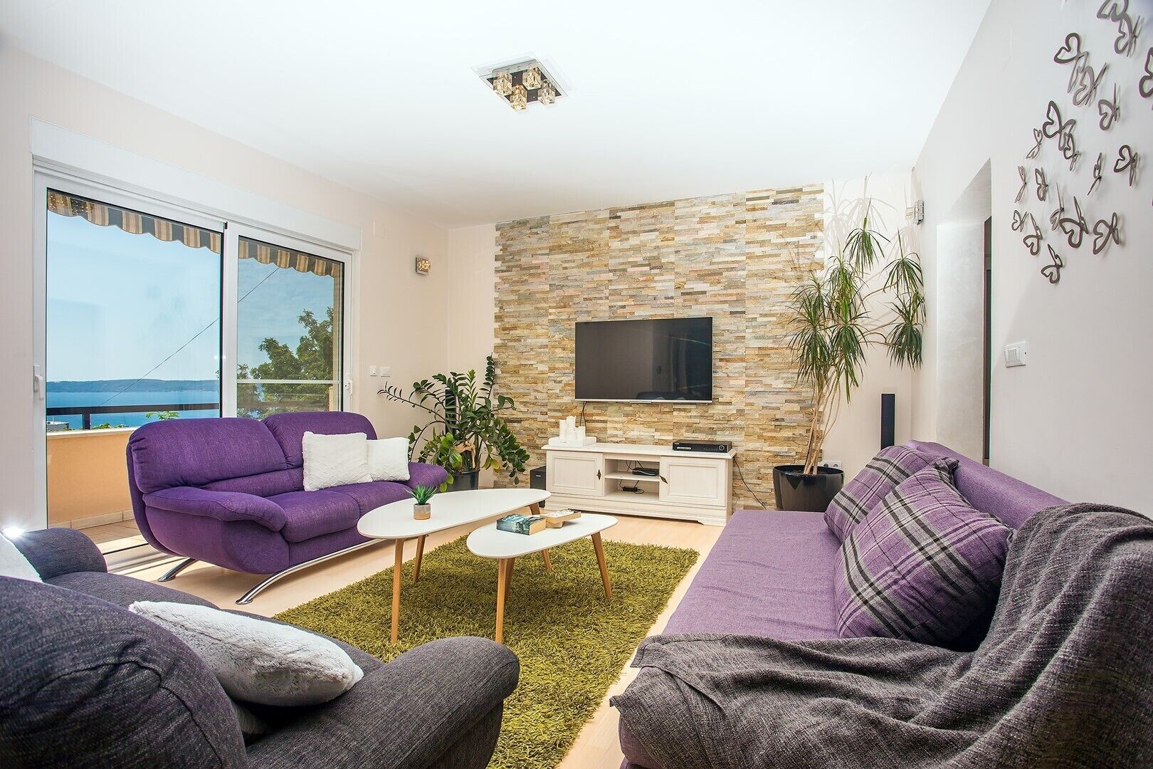 Ein modernes Wohnzimmer mit einer Wand aus Stein, einem Flachbildfernseher und Meerblick durch große Fenster.