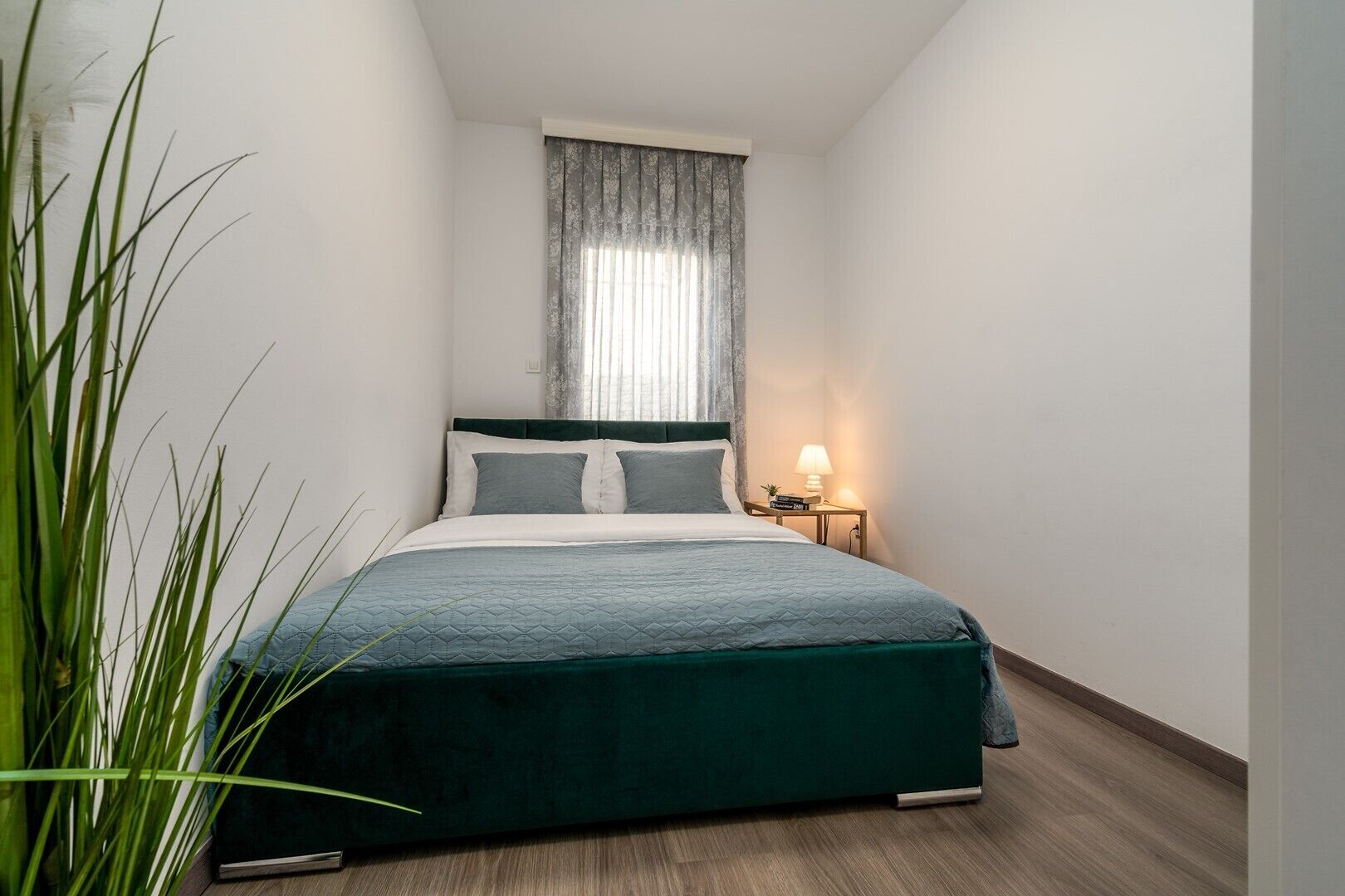 A narrow hotel room with a bed, a bedside table with a lamp, and a window with curtains.