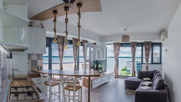 Une cuisine moderne avec un îlot central en bois, un coin repas offrant une vue sur la mer et un salon avec un canapé et un téléviseur.