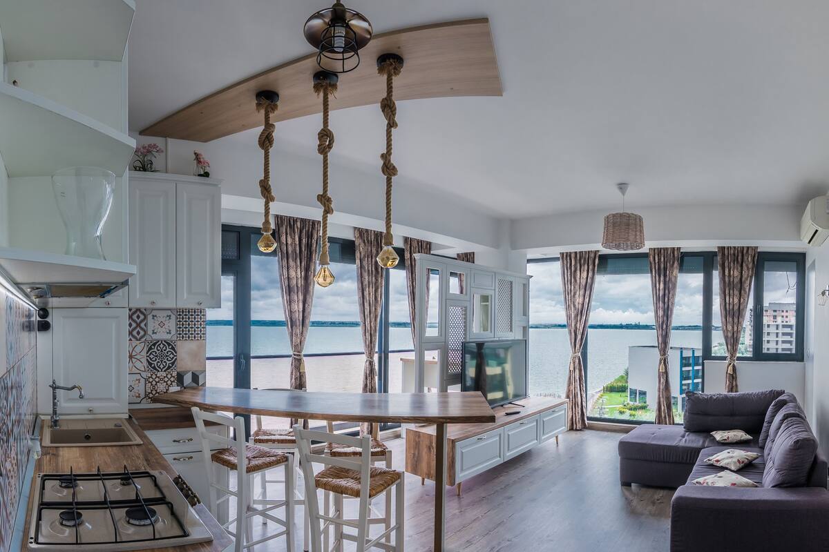 A modern kitchen with a wooden island, a dining area with a view of the sea, and a living room with a sofa and a TV.