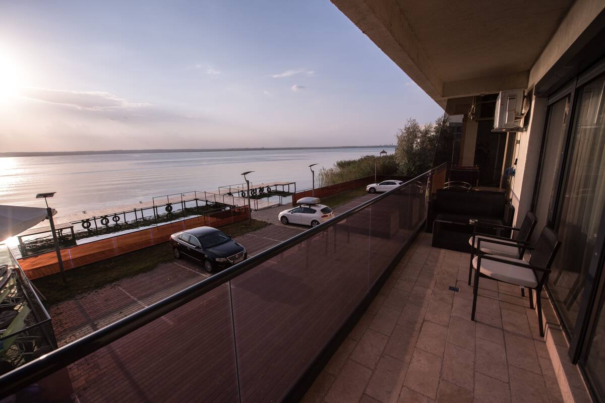 A balcony with a view of the sea, a carport with parked cars, and outdoor furniture.