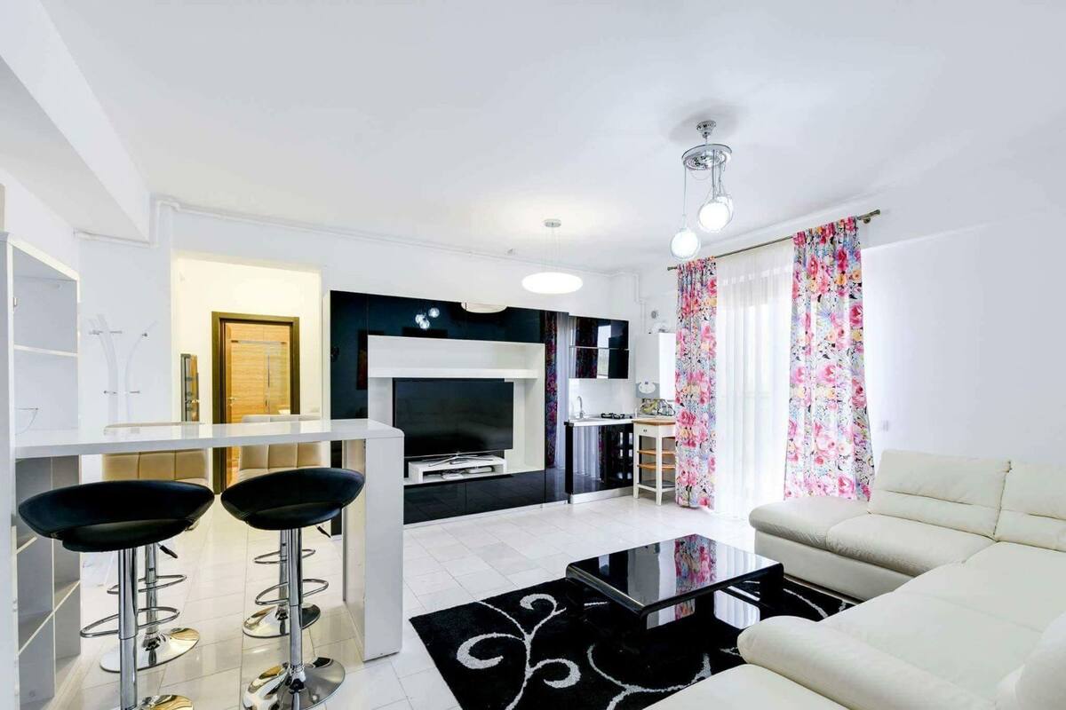 A modern living room with a sofa, a coffee table, and two bar stools. There is a fireplace, a television, and a kitchen area in the background.