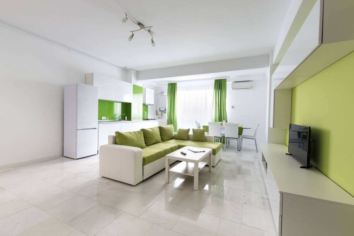A modern living room with a white sofa, a coffee table, and a TV. There is a kitchen area with white cabinets and a green countertop.