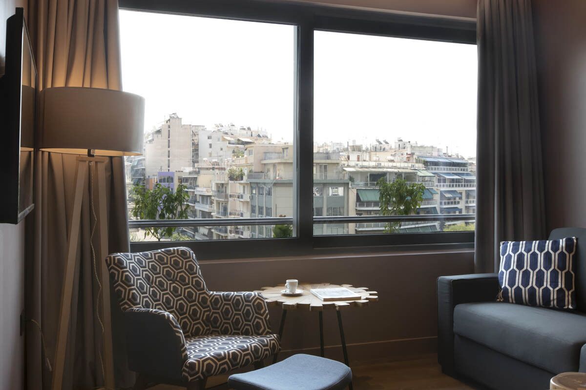 A modern hotel room with a large window offering a city view, a patterned armchair, a small table with a cup, and a sofa with a patterned pillow.