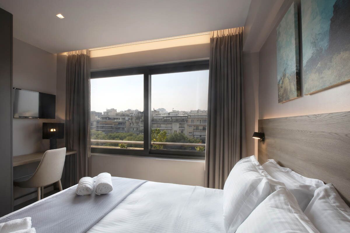 A hotel room with a large window, a bed with white linens, a desk with a chair, and a wall-mounted TV.