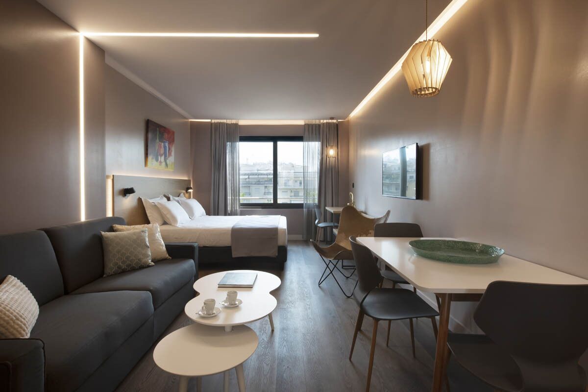 A modern hotel room with a bed, a sofa, a dining area, and a view of the city.