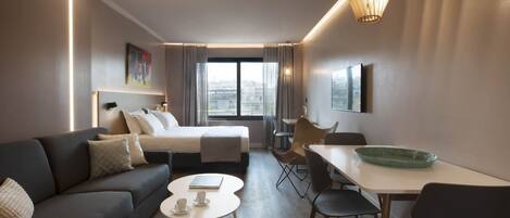 A modern hotel room with a bed, a sofa, a dining area, and a view of the city.