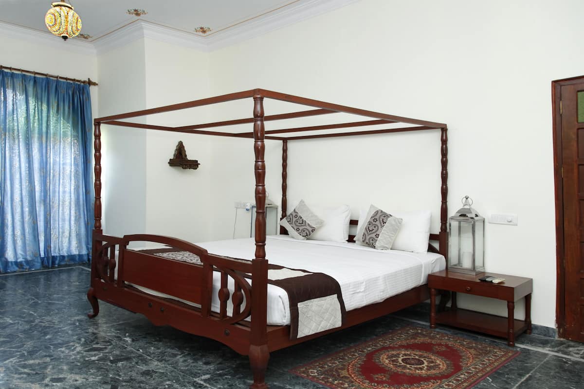 A four-poster bed with a wooden frame, white bedding, and patterned pillows. There is a nightstand with a lamp and a decorative item beside the bed. The room has a marble floor and a red patterned rug.