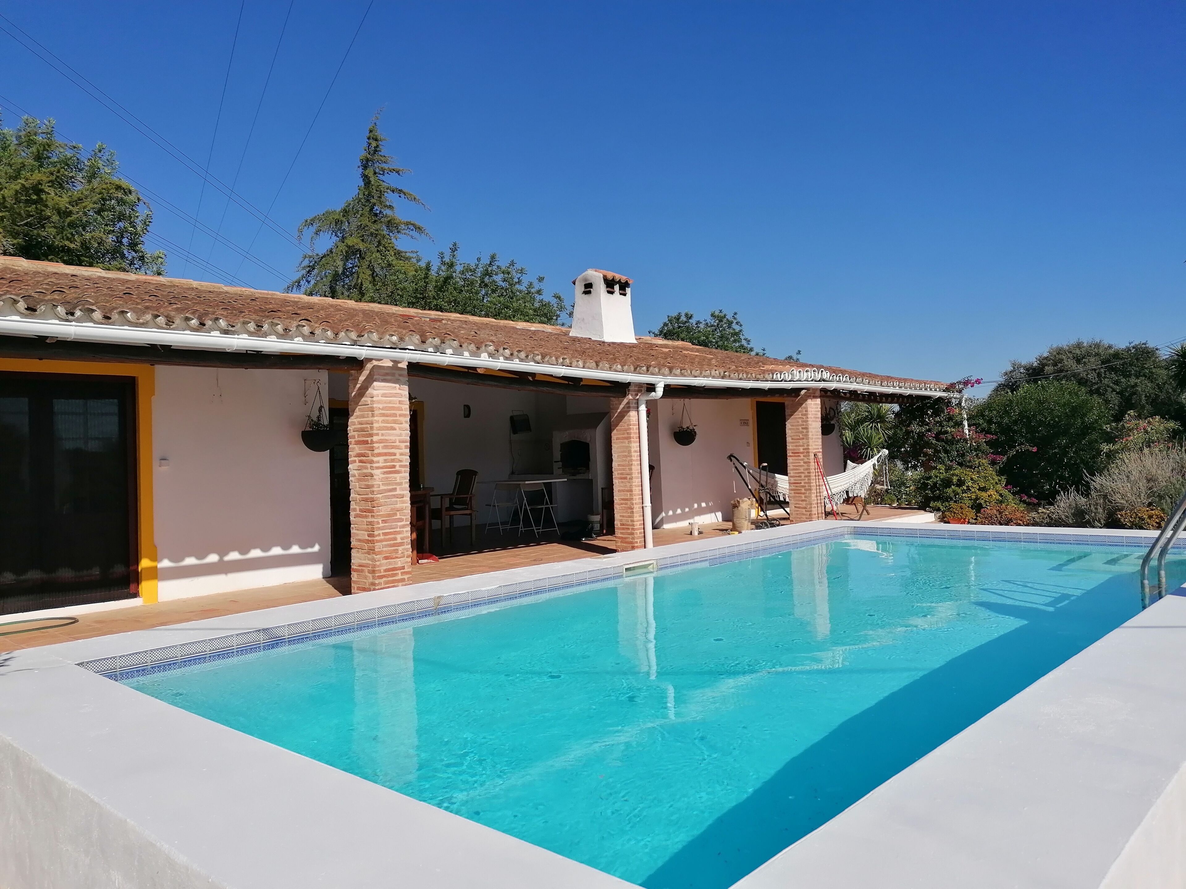 A swimming pool with a tiled edge, a covered patio area with outdoor furniture, and a brick building with a chimney.