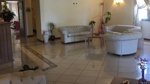 A spacious living room with a large sofa, a smaller armchair, and a coffee table. There is a potted plant and a lamp on the table. The room has a tiled floor and a ceiling with recessed lighting. - Hotel Terra di Gallura (Budoni)