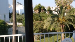 Un balcon doté d’une balustrade blanche, donnant sur un jardin planté de palmiers et d’autres végétaux, avec des bâtiments en arrière-plan.