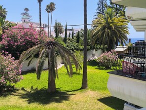 Un jardin bien entretenu, agrémenté de palmiers et d’arbustes en fleurs, un sentier et un balcon avec du mobilier d’extérieur.