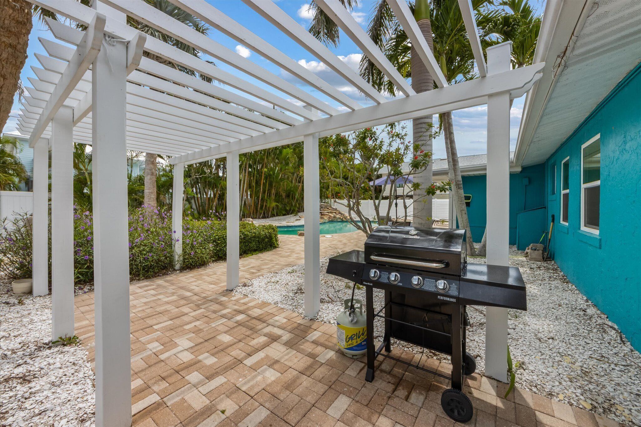 Un patio couvert avec un barbecue, une pergola blanche et une piscine en arrière-plan.