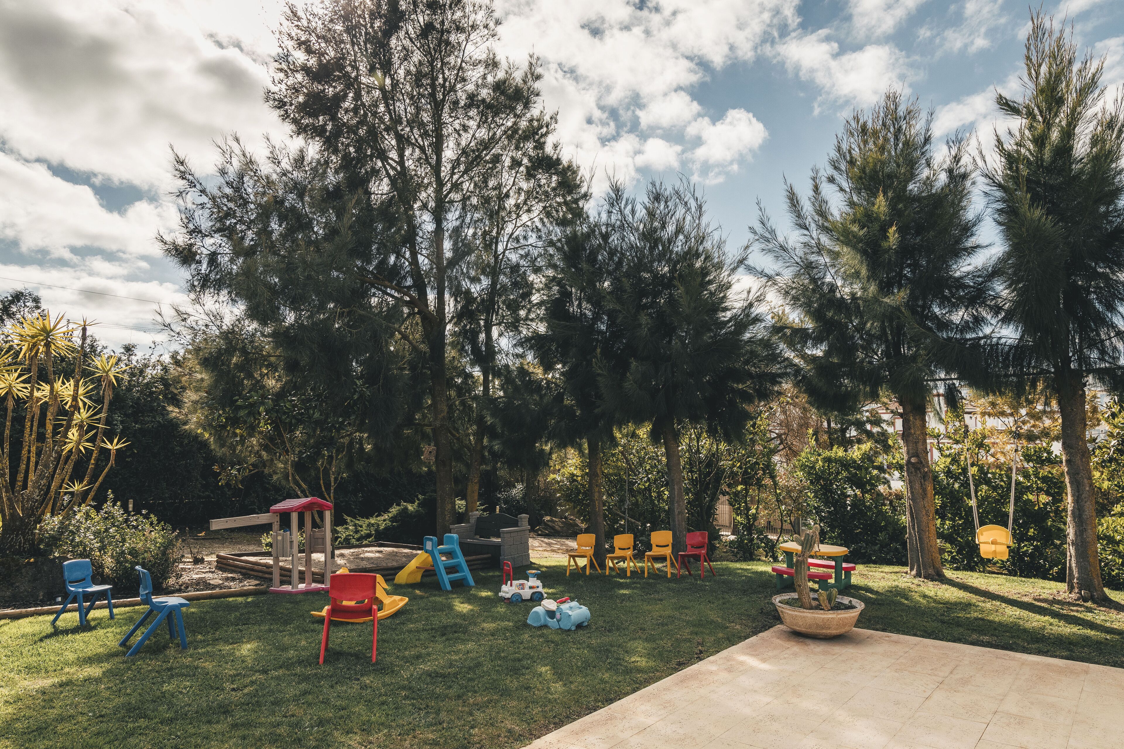 Ein Garten mit Spielbereich, darunter eine Rutsche und Schaukeln, umgeben von Bäumen und blauem Himmel.