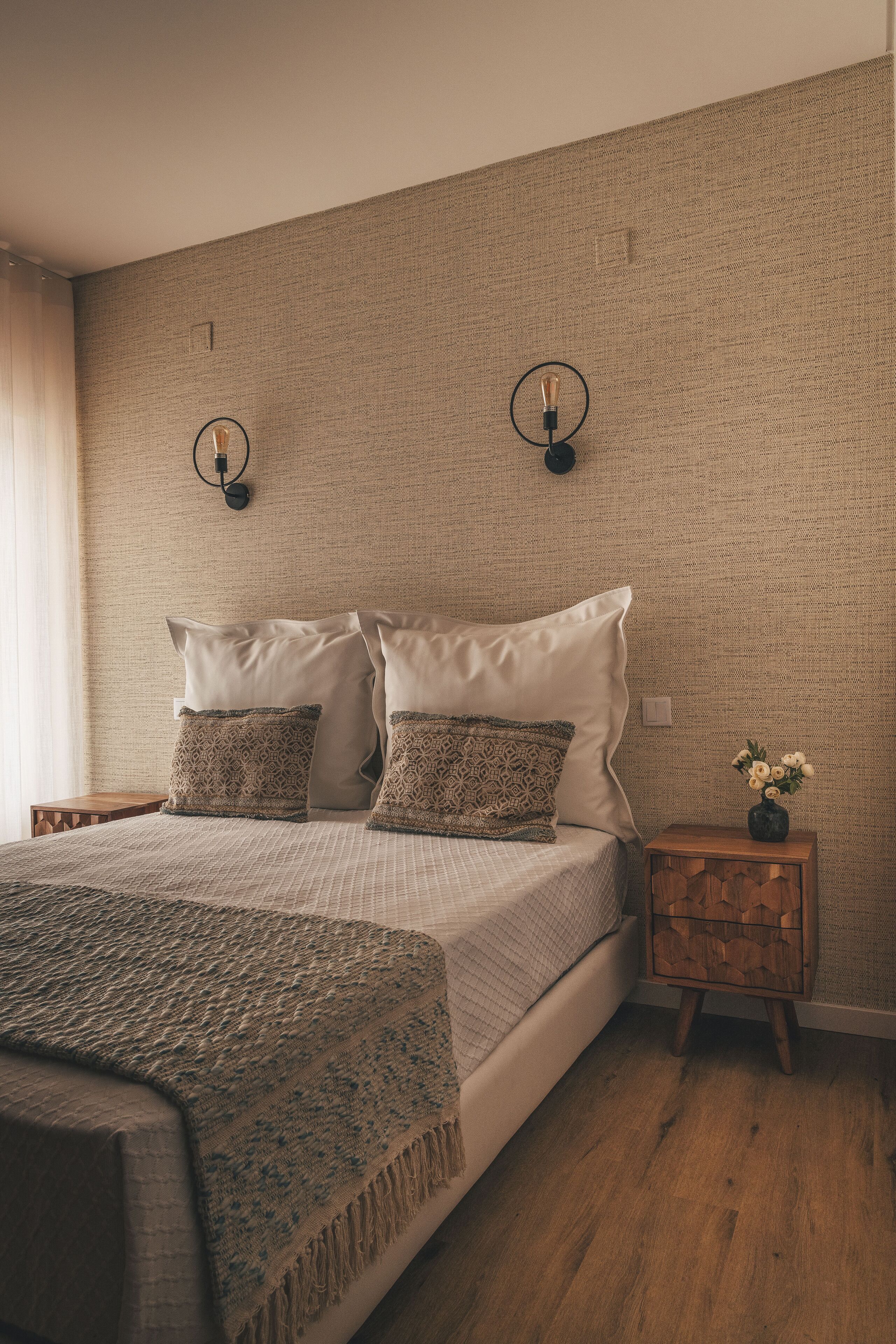 A neatly made bed with patterned pillows, a wooden nightstand with a vase of flowers, and a textured wall.