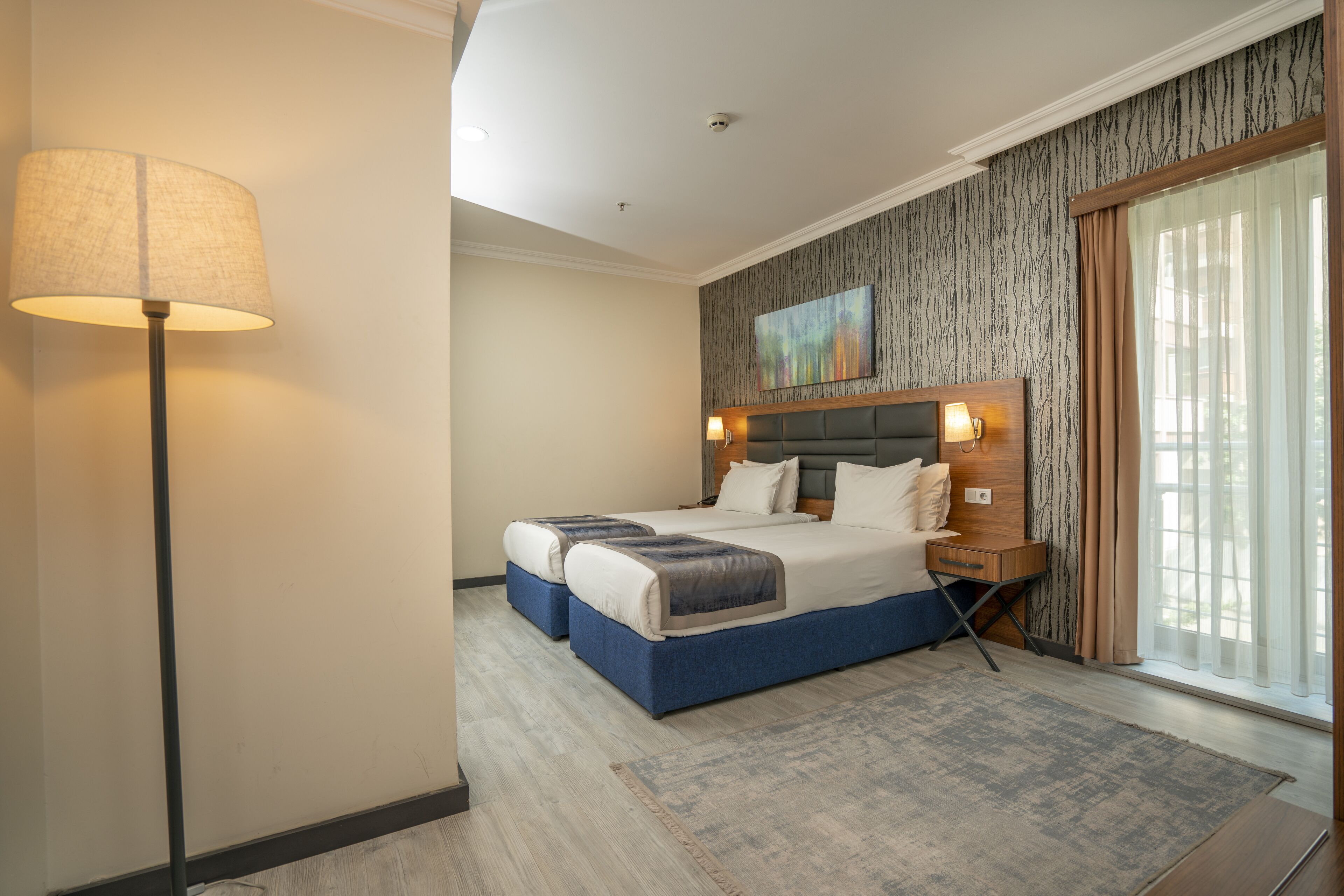 A hotel room with two beds, a large floor lamp, a painting on the wall, and a view of the outside through a window.