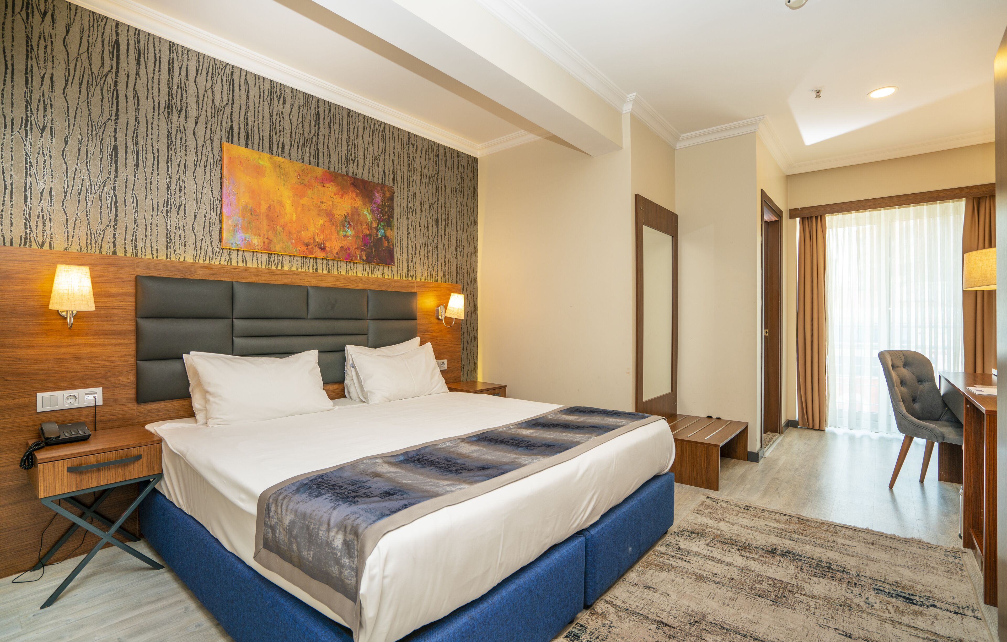 A hotel room with a large bed, a wooden headboard, a wall-mounted artwork, two bedside lamps, a desk with a chair, and a large mirror.